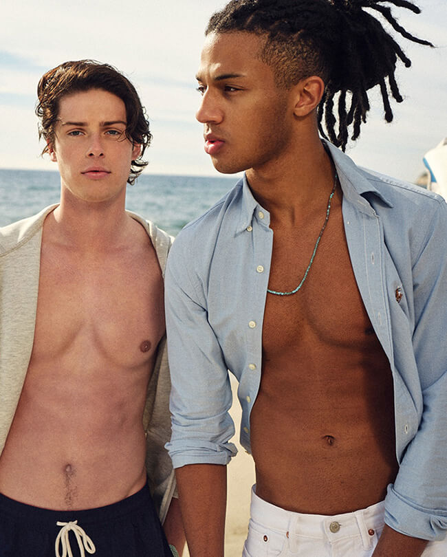 Two male models on beach wearing shorts and open shirts showing their well chiseled abs.